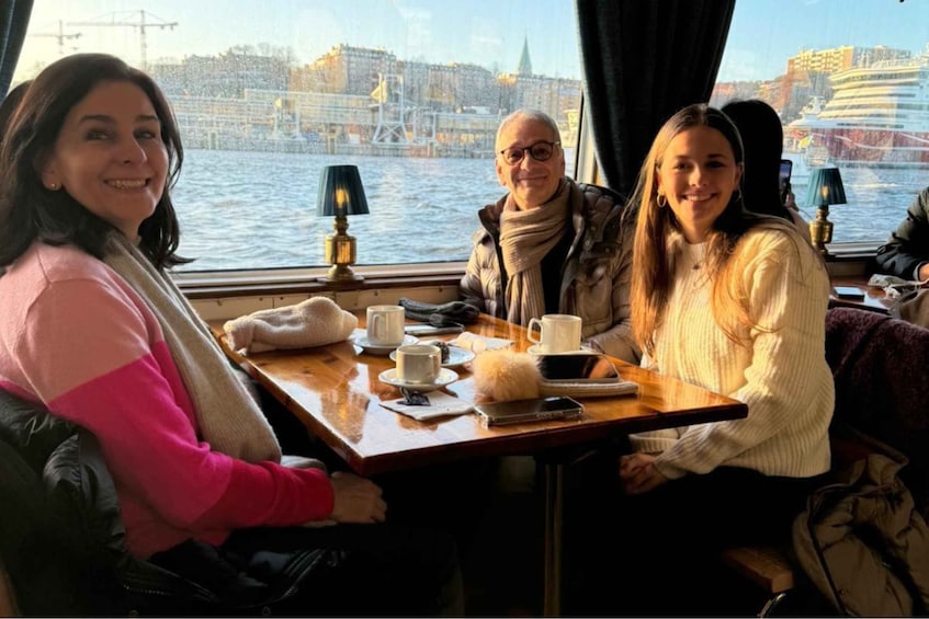 Three people seated at a table with a view of a river and buildings in the background. 1