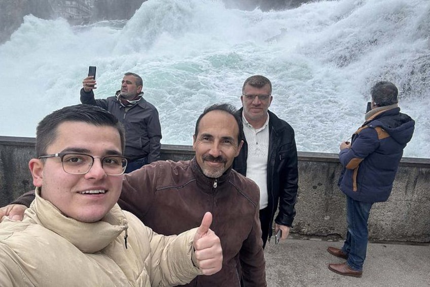 A group of people standing by a large, turbulent sea with waves crashing against a concrete barrier. 4
