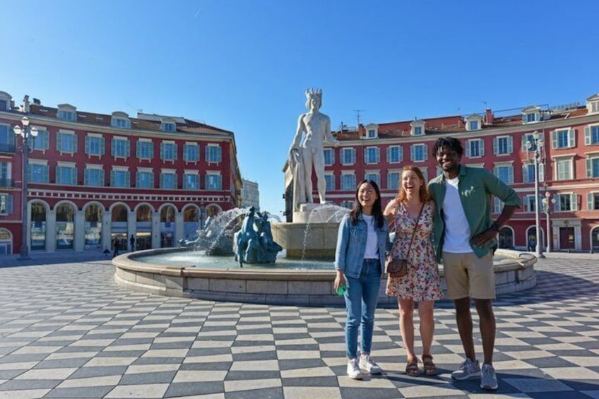 A plaza with a fountain, a statue, and red buildings with arched windows. 1