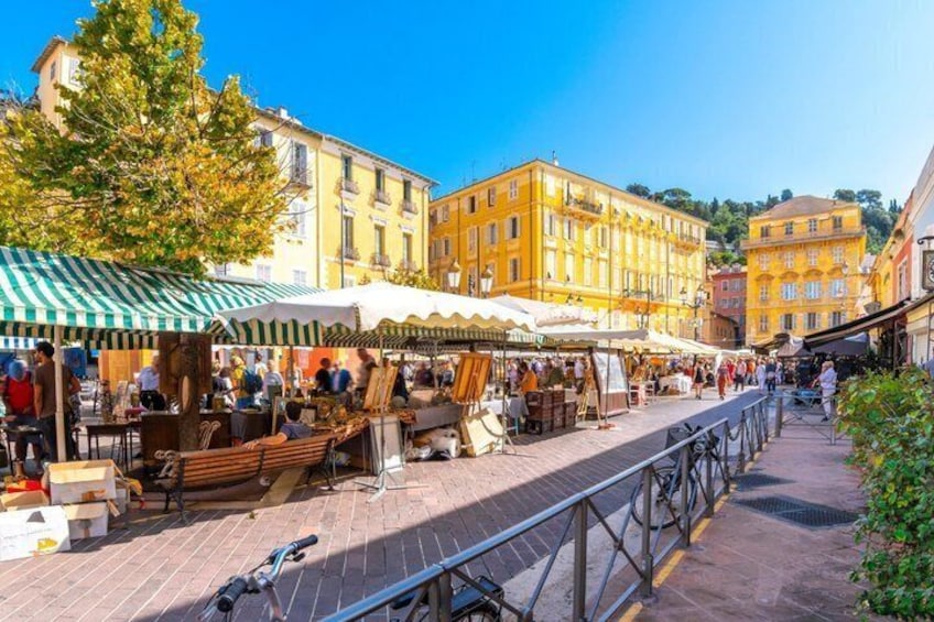 A bustling outdoor market with vendors and stalls, set against a backdrop of historic yellow buildings and a clear blue sky. 5