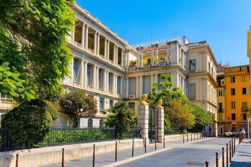 A classical building with columns, a paved sidewalk, and a fence. 2