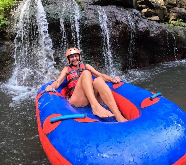 Eine Person sitzt in einem aufblasbaren blauen und roten Schlauchboot auf einem Fluss, im Hintergrund ein Wasserfall. 5