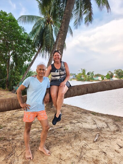 Two people sitting on a tree branch by a sandy beach with palm trees and a body of water in the background. 2