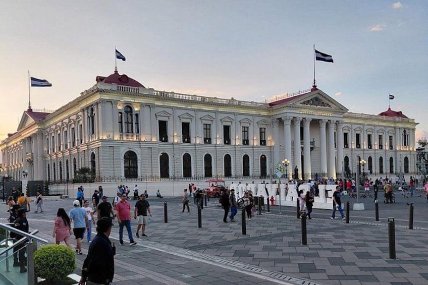 A large, classical building with columns and flags, surrounded by a busy public square with people walking and gathering. 5