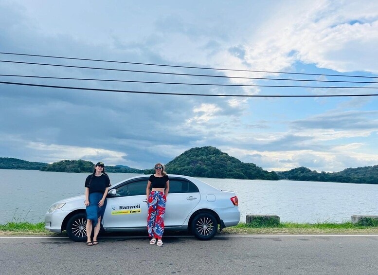 Two people standing beside a white car with 'Ranwel Tours' on the side, parked by a lake with hills in the background. 2