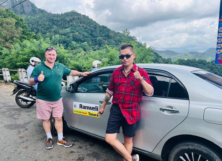 Two people standing by a silver car with a 'Ranwel Tours' sign, a motorcycle, and a mountainous landscape in the background. 1