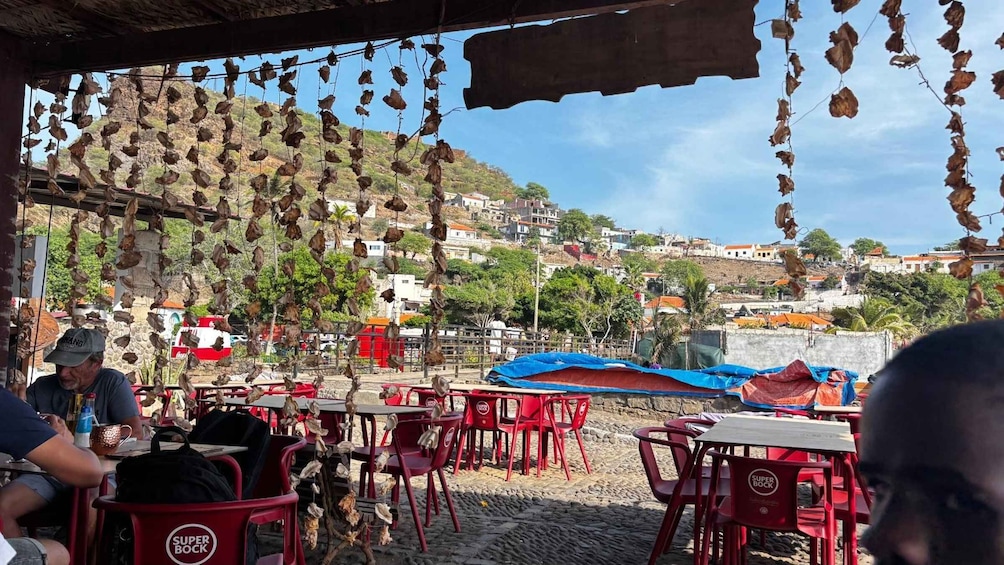 A coastal restaurant with red tables and chairs, hanging dried fish, and a view of a hillside town. 4