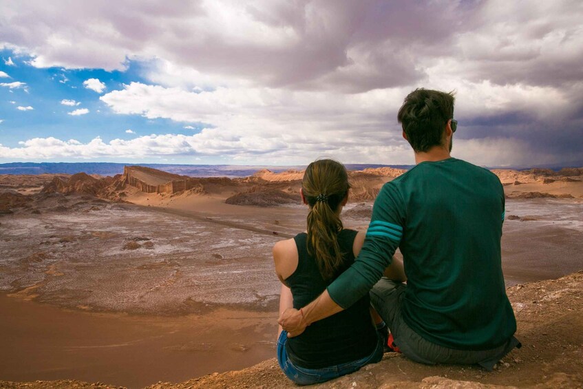 Dos personas sentadas sobre un afloramiento rocoso, con vista a un paisaje árido y colinas distantes. 2