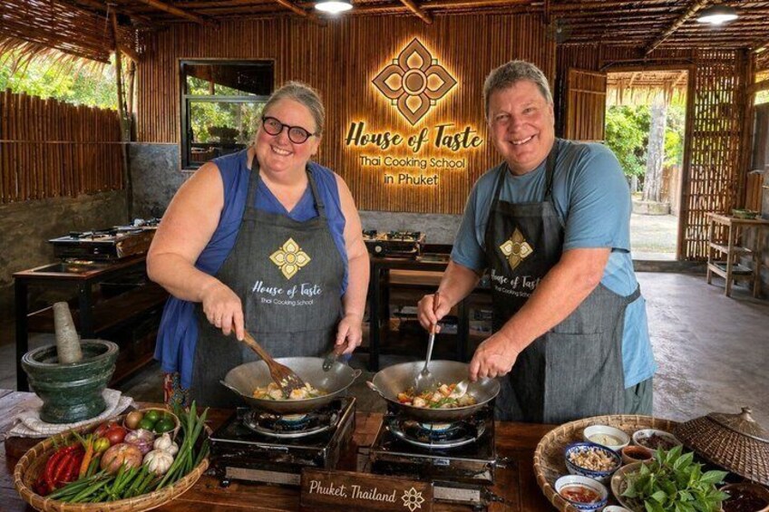 Two people cooking in a kitchen with a sign that reads 'House of Taste Thai Cooking School in Phuket.' 1