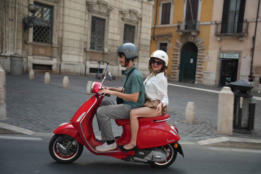 A red scooter with two passengers, a man and a woman, on a cobblestone street with historic buildings in the background. 5