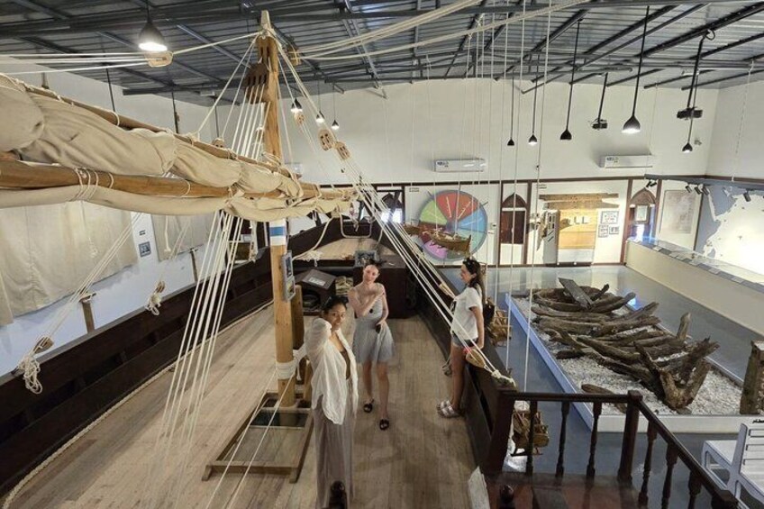 A museum exhibit with a large sailing rigging structure, a display case with artifacts, and a wooden floor. 3
