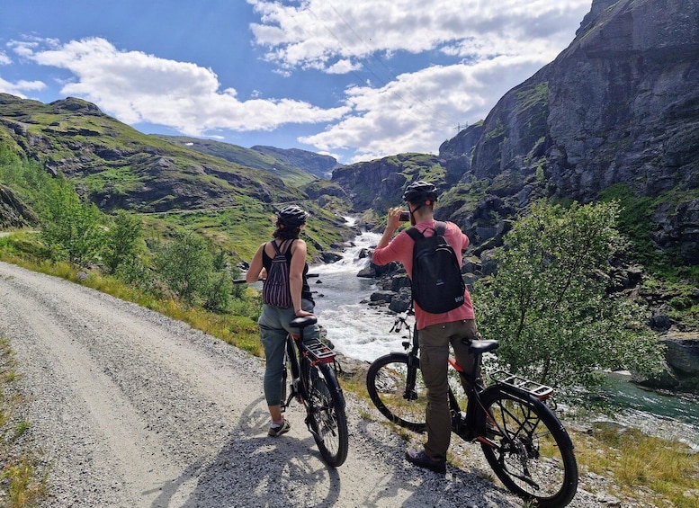 Two cyclists on a gravel road overlooking a river with a mountainous backdrop. 5