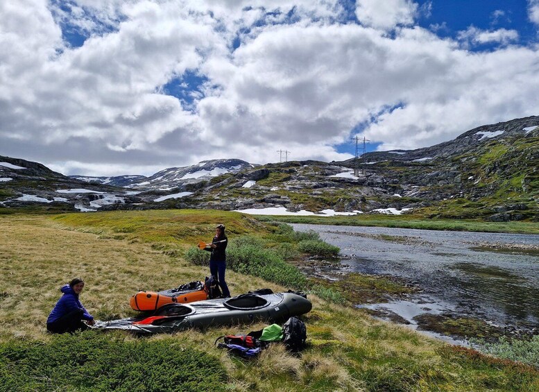 A kayaker preparing on a grassy riverbank with mountains and power lines in the background. 3