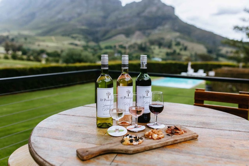 A wooden table with three wine bottles, glasses, and a wooden board with snacks, set against a mountainous backdrop. 2