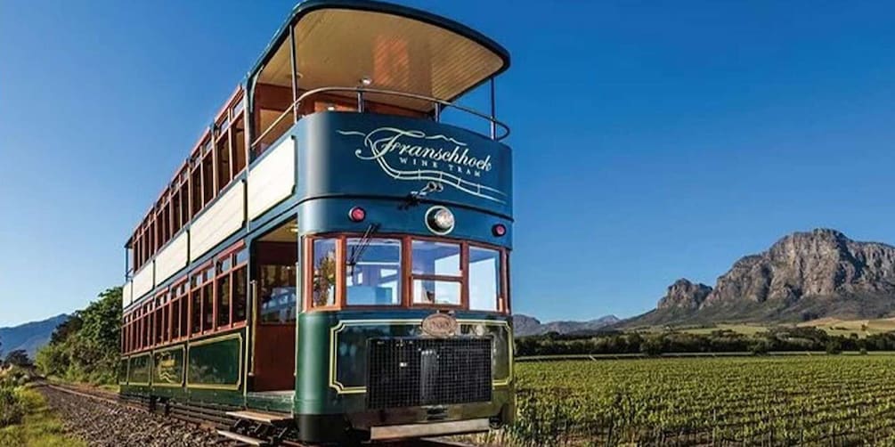 A vintage tram with 'Franschhoek Wine Tram' written on it, traveling on tracks with a vineyard and mountains in the background. 1