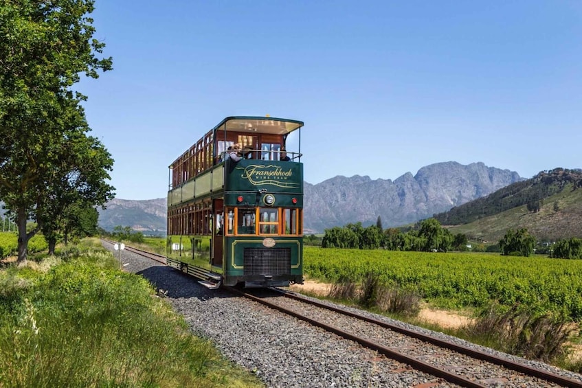 A green double-decker train with 'Franschhoek Wine Tram' written on it, traveling on tracks surrounded by vineyards and mountains. 5