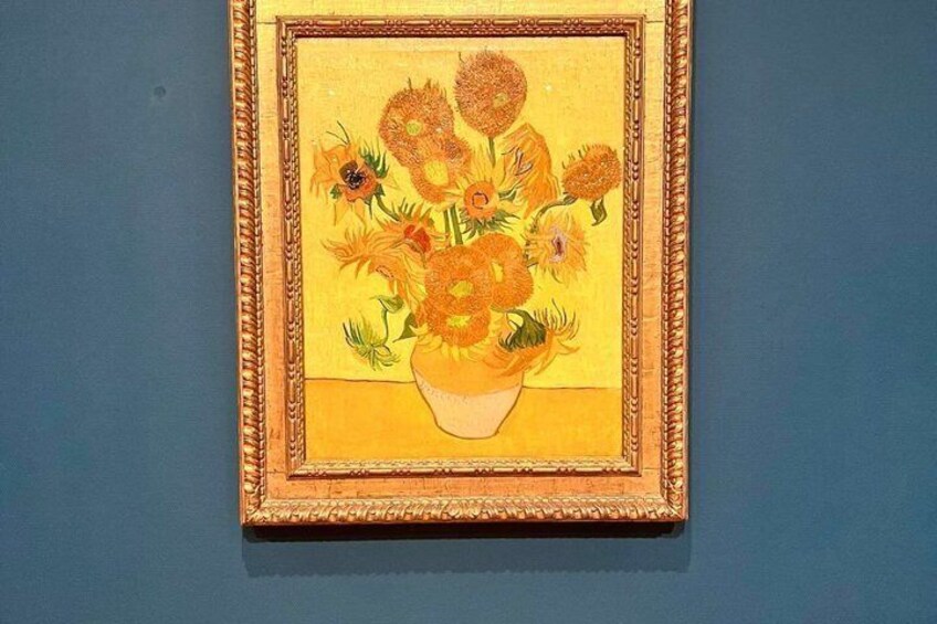 A framed painting of sunflowers in a vase on a blue wall. 2