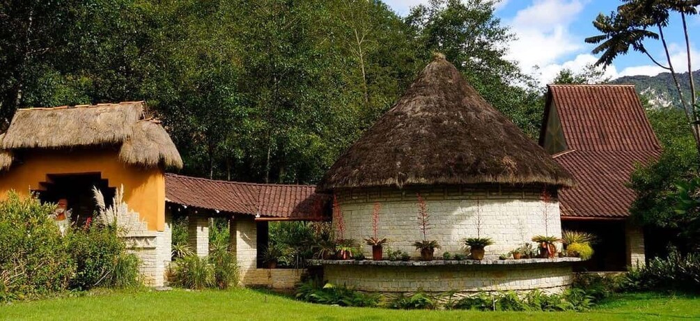 A traditional thatched-roof house with a white wall and a red-tiled roof, surrounded by lush greenery and a clear sky. 1