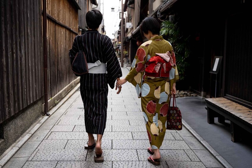Two individuals walking down a narrow alleyway, one in a striped kimono and the other in a kimono with a floral pattern, holding hands. 3
