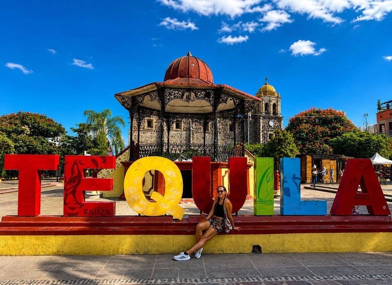 A person sitting on a colorful bench with the word 'TEQUILA' in large letters, a gazebo, and a historic building in the background. 1