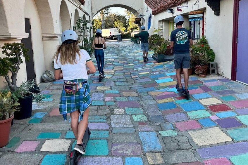 Una calle adoquinada y colorida con peatones y una persona montando un patinete. 1