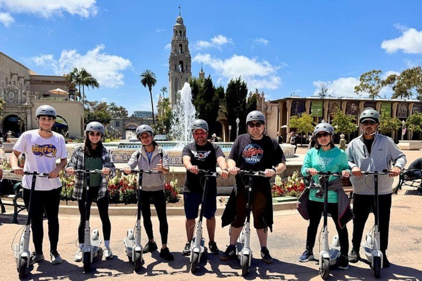 A group of people on electric scooters in an outdoor setting with a fountain and a bell tower in the background. 3