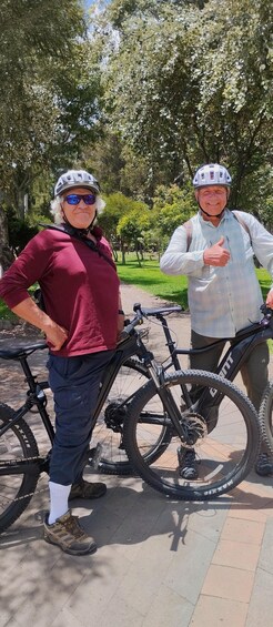 Two people standing next to bicycles in a park with trees and a paved path. 1