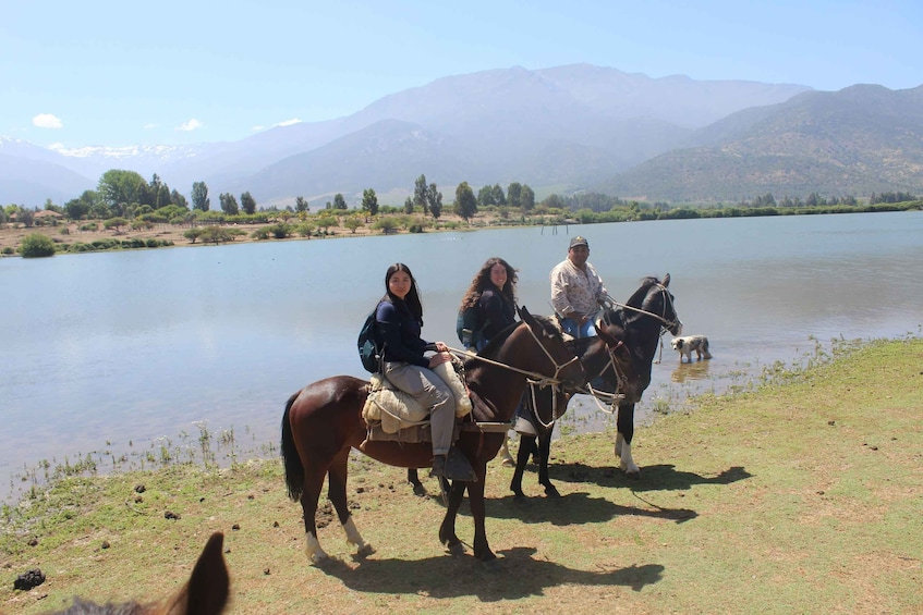 Three people on horseback by a lake with mountains in the background. 2