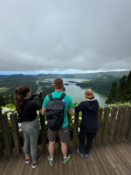 Three people standing on a wooden deck overlooking a valley with a lake and mountains. 2