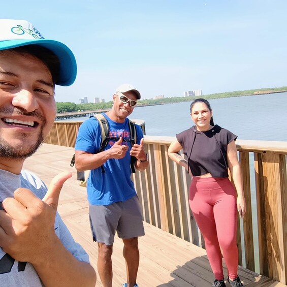 Three people on a wooden boardwalk by a river with buildings in the background. 1