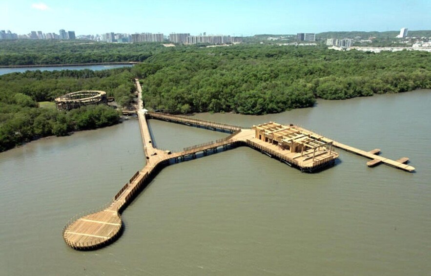 A large, circular wooden structure in the water, surrounded by dense greenery and a distant cityscape. 5