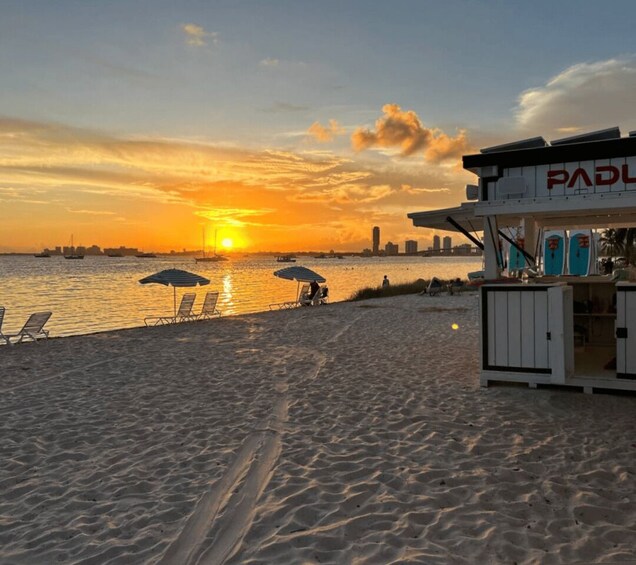 A beach with lounge chairs and umbrellas, a paddleboard rental shack, and a sunset over the water. 1