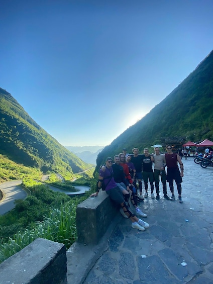 A group of people sitting and standing on a stone ledge overlooking a valley with a winding road and a river. 2