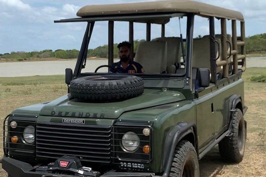 A green Defender vehicle with a spare tire mounted on the front, parked on a dirt road with a body of water and greenery in the background. 3