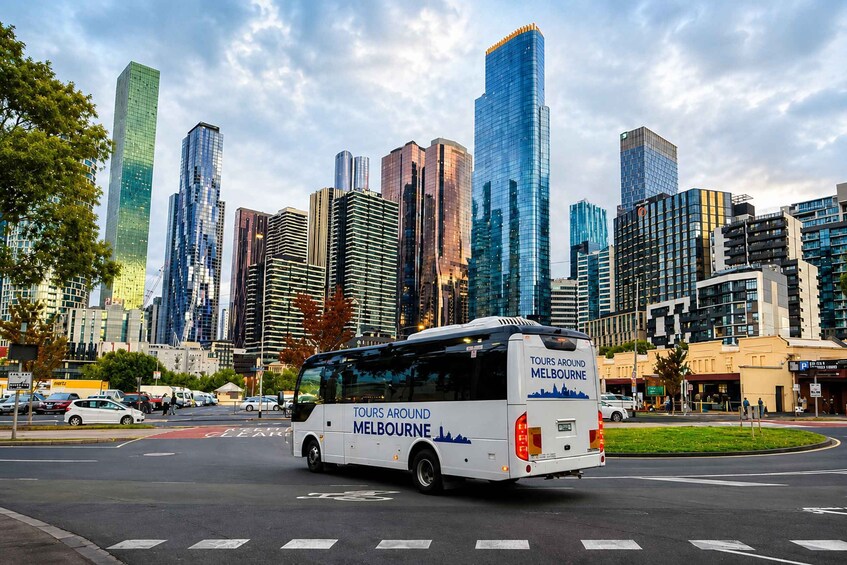 A cityscape with modern skyscrapers and a tour bus labeled 'TOURS AROUND MELBOURNE' driving on a road. 1