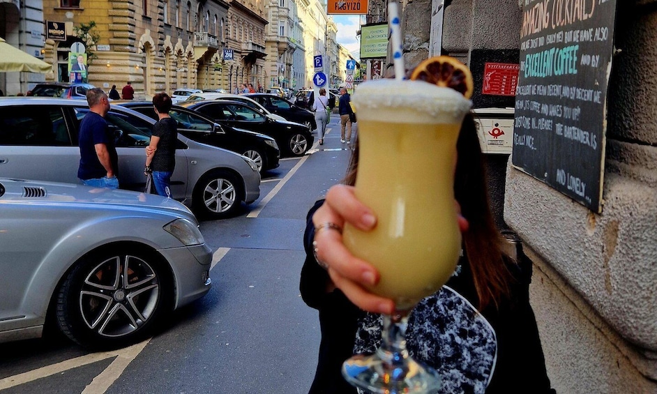 A person holding a large glass of beer in an urban street scene with cars and a building with a sign. 5