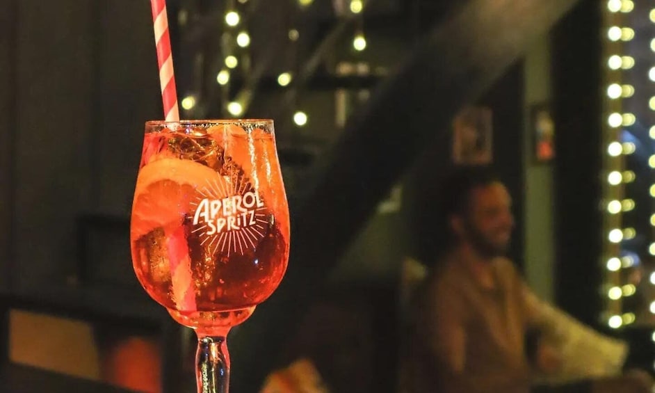 A glass of a red beverage with a straw, labeled 'Aperol Spritz,' in a dimly lit bar setting. 3