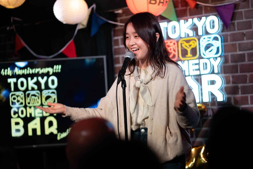 A person speaking into a microphone with a neon sign in the background that reads 'TOKYO COMEDY BAR.' 2