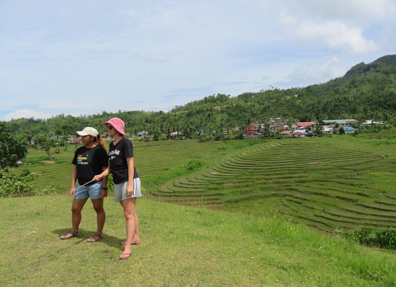Two people standing on a grassy hill with terraced fields and houses in the background. 4
