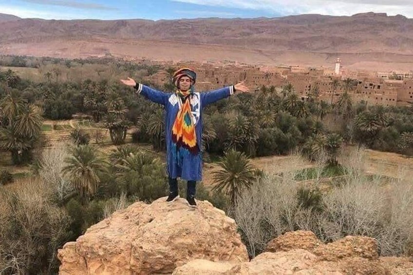 A person standing on a rock with arms outstretched, palm trees, and a desert landscape with buildings in the background. 4