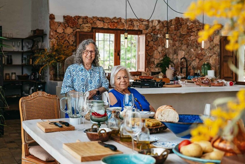 Two people seated at a table with food and drinks, in a kitchen with stone walls and wooden furniture. 1