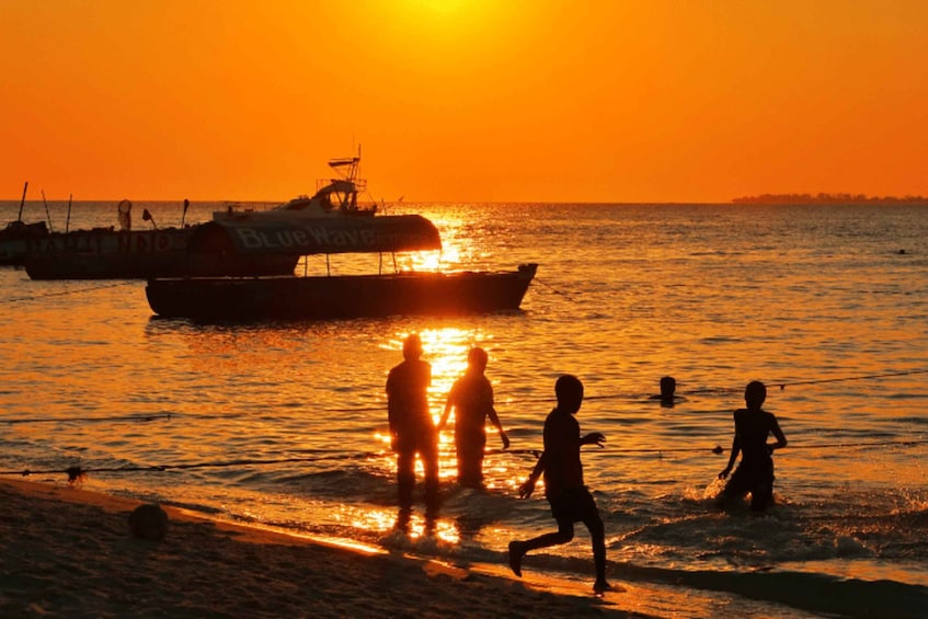 Un tramonto su una spiaggia con persone che giocano in acqua e una barca chiamata 'Blue Wave' sullo sfondo. 1