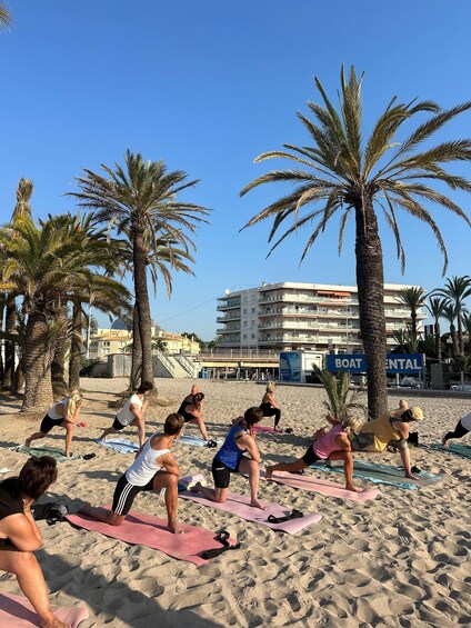 A group of people doing yoga on mats on a beach with palm trees and a boat rental sign in the background. 2