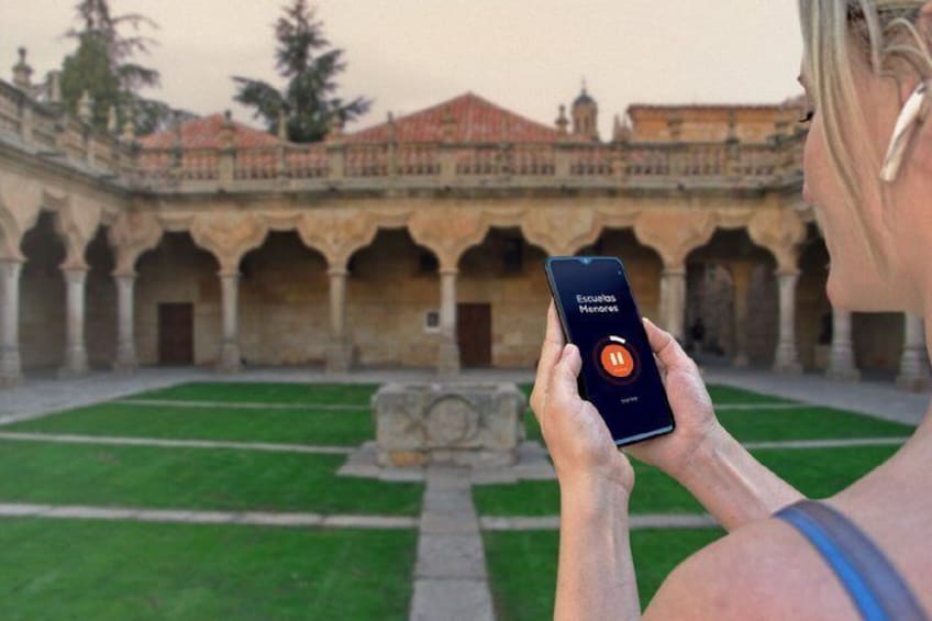 A person holding a smartphone with a music app open, standing in a courtyard with a fountain and a building with arches in the background. 5