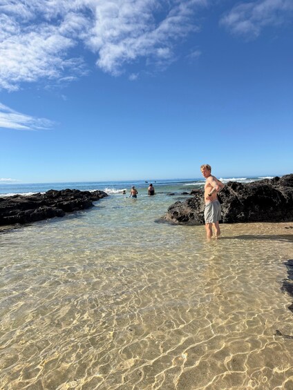 A beach with clear shallow waters, rocky shore, and a person standing near the water's edge. 4