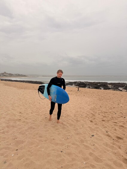 A person in a black wetsuit carrying a blue surfboard on a sandy beach with rocky outcrops in the background. 2