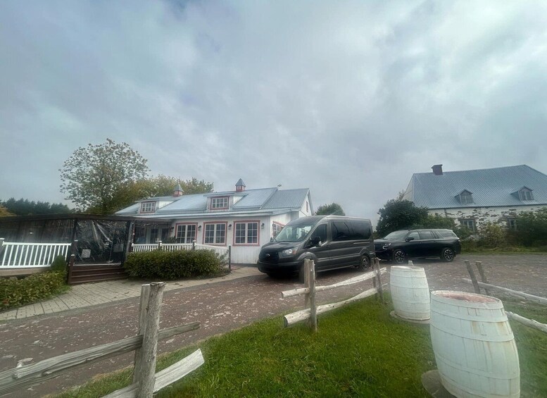 A rural setting with a white building with a red trim, a black van, and a black car parked in front. There are wooden barrels and a wooden fence in the foreground. 3
