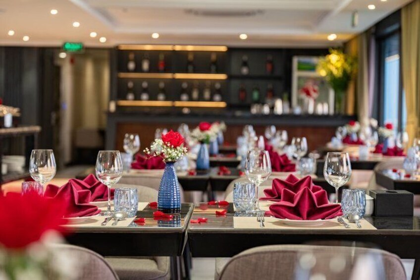 Un interno di ristorante con tavoli apparecchiati per la cena, con tovaglioli rossi, bicchieri trasparenti e vasi azzurri con fiori. 2