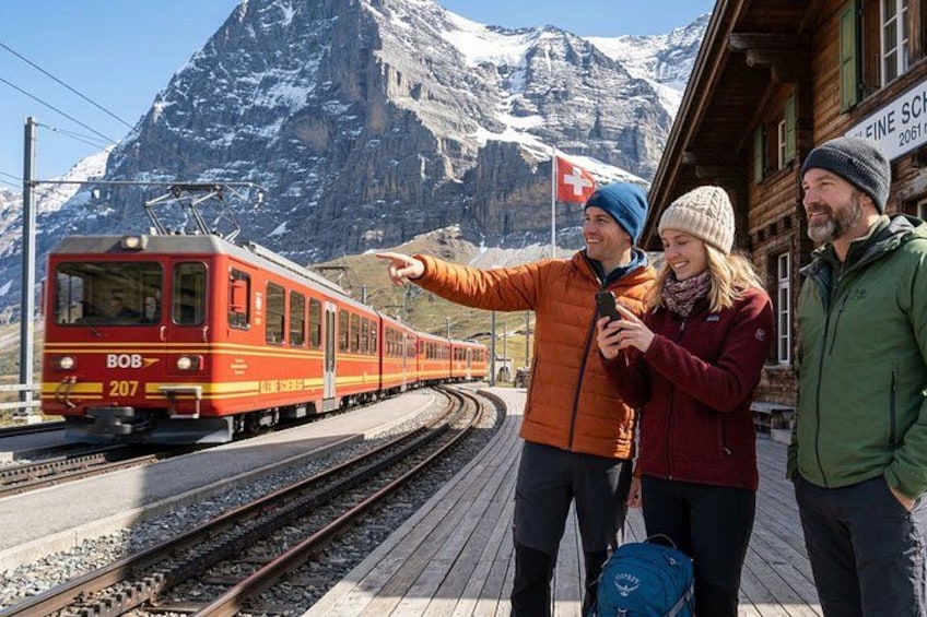 A red train on tracks with a mountainous background, a wooden building, and people on a platform. 5