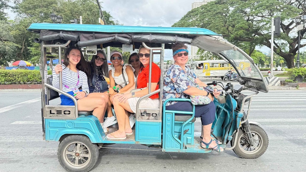 A group of people on a teal tuk-tuk with 'LEO' branding, parked on a city street with trees and buildings in the background. 3